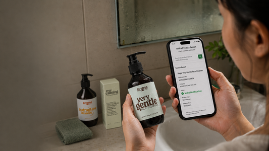 A person in a bathroom holds a smartphone showing a NPRA product app screen while holding a dark amber bottle labeled 'very gentle' by SEGAR. Other SEGAR products and a sponge sit on the bathroom counter in the background.